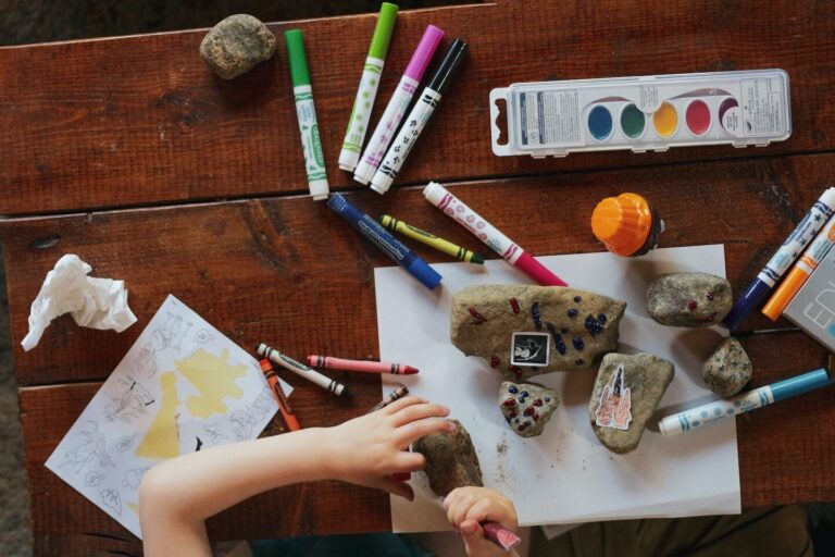 A child painting rocks at a wooden table covered with markers, crayons and watercolors